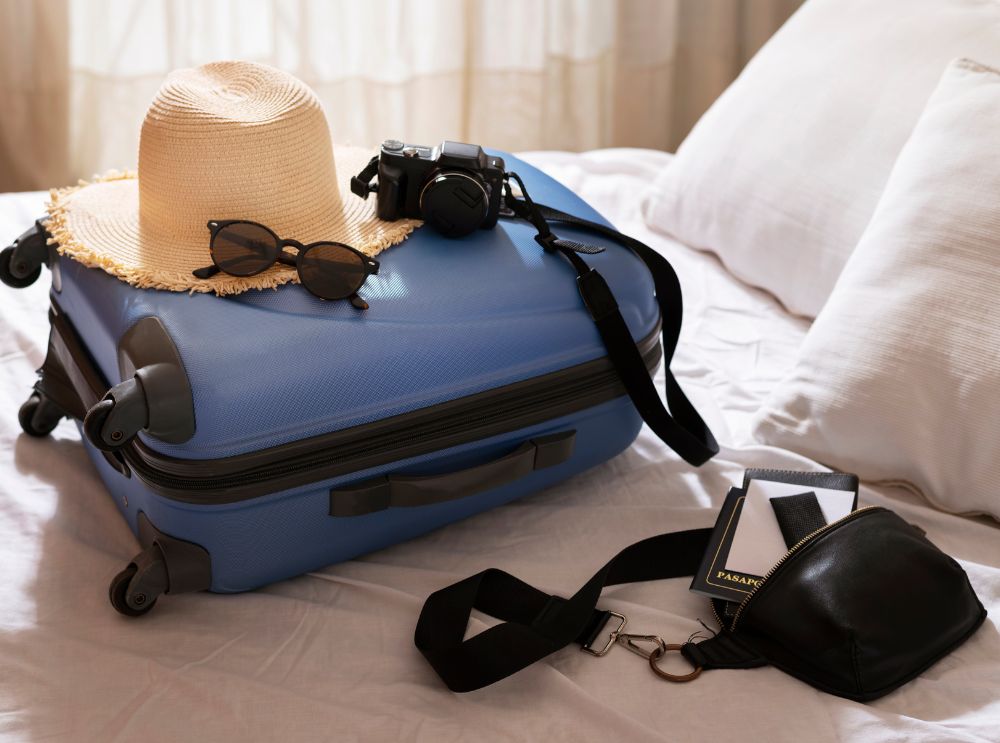 Uma mala azul com um chapéu de palha, óculos de sol e uma câmera fotográfica descansam em uma cama bem arrumada, acompanhados de um passaporte e uma pochete. Ao fundo, uma cortina deixa entrar luz suave no ambiente.