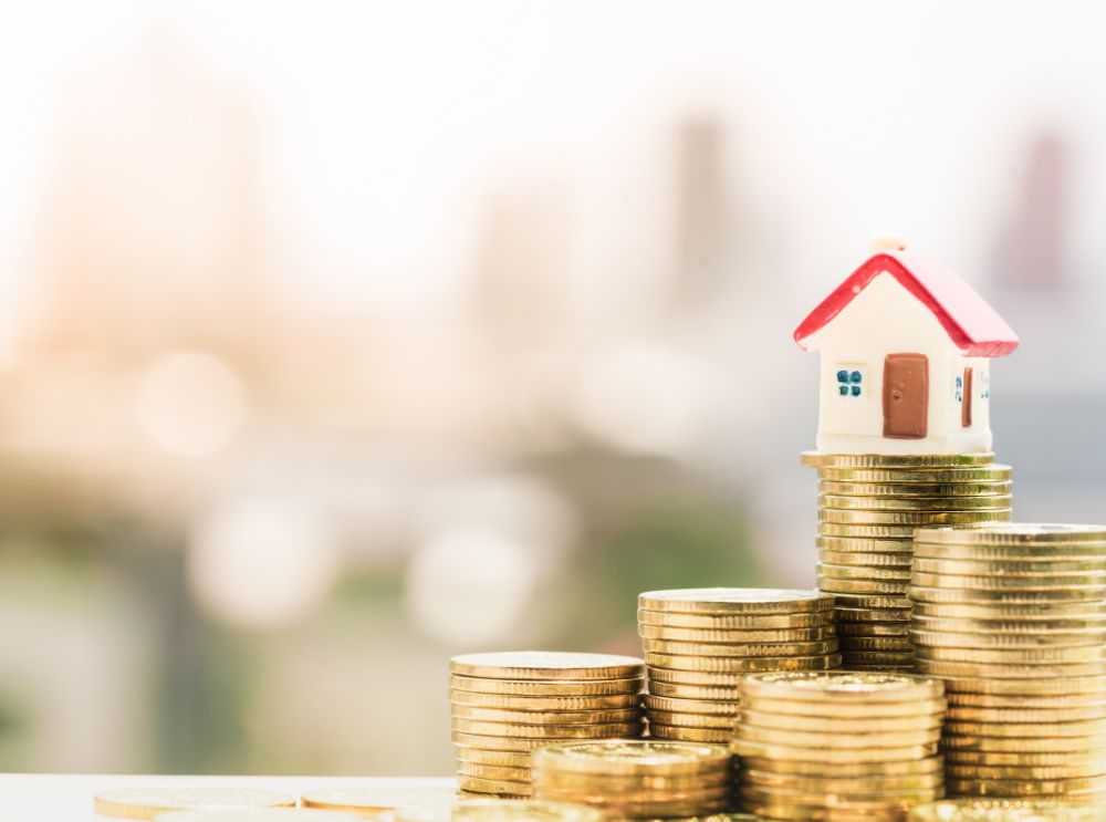 Pequena casa em miniatura com telhado vermelho sobre pilhas de moedas douradas, representando investimento imobiliário. O fundo desfocado mostra uma paisagem urbana iluminada.