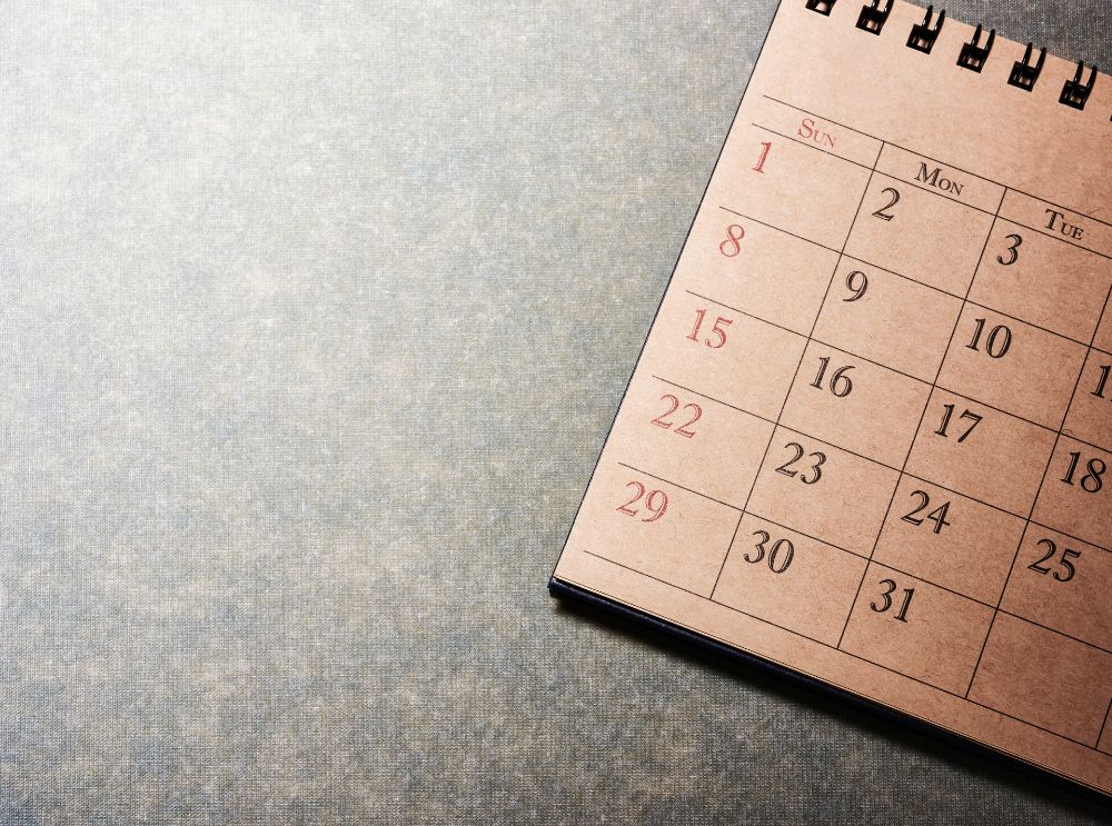 Calendário de papel com fundo bege, posicionado sobre uma superfície texturizada cinza. Os números dos dias estão impressos em preto, com os domingos destacados em vermelho.
