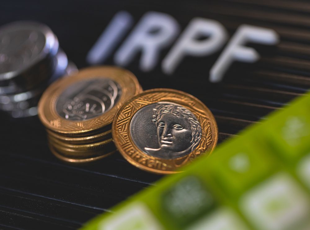 Moedas de real empilhadas sobre um teclado preto, com a sigla "IRPF" em destaque ao fundo e parte de um teclado numérico verde em primeiro plano.