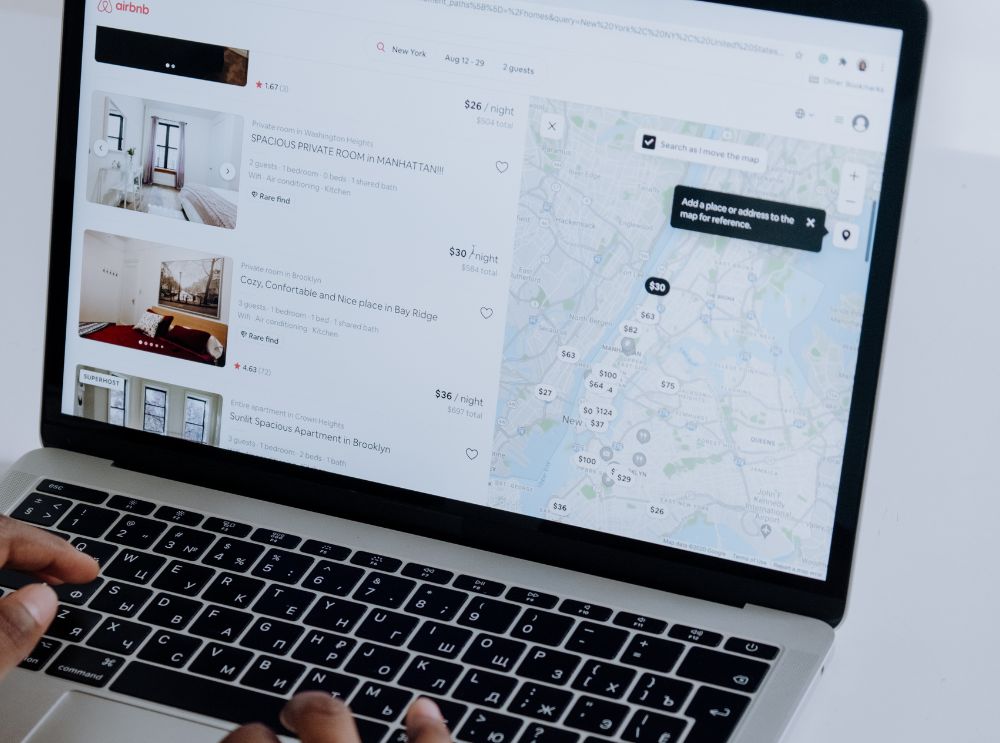 Pessoa usando um laptop com a tela exibindo o site do Airbnb. A página mostra opções de acomodações com fotos e preços à esquerda, e um mapa com localizações e valores à direita. As mãos do usuário estão visíveis no teclado.