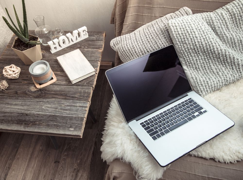Notebook prata sobre uma manta felpuda em um sofá com almofadas cinzas, ao lado de uma mesa de madeira com objetos decorativos e planta em vaso.