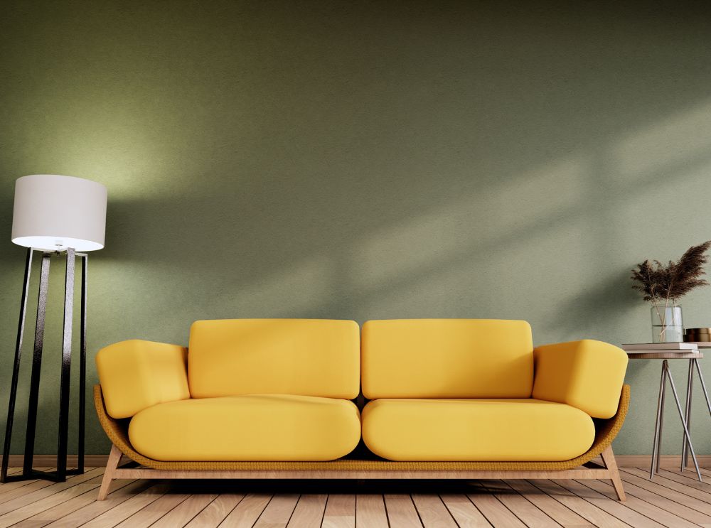 Sofá amarelo com design moderno em madeira clara, posicionado entre luminária de chão preta e mesa lateral com vaso de plantas secas, sobre piso de madeira clara e parede verde.